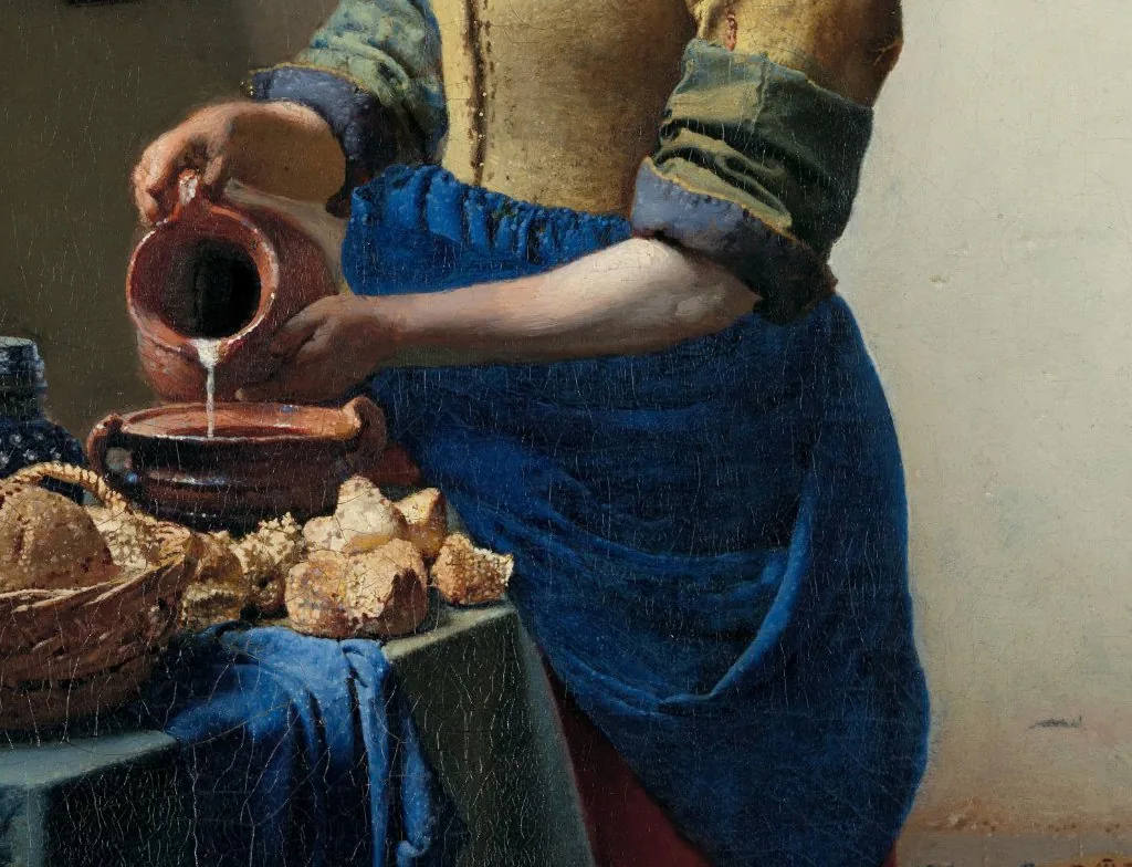 A detail of the same Milkmaid photograph, cropped toward the table: bread, a white jug, the stream of milk, a yellow bodice, and a blue apron.
